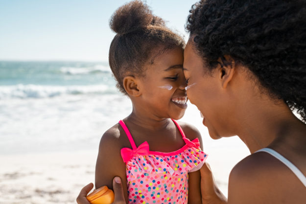 Woman hugging lovely daughter with sunscreen on the cheeks Young black mother embracing daughter with sunscreen on face at beach with copy space. Woman with closed eyes face to face with her cute little girl with sun lotion on face. Funny black female kid with sunblock at seaside with her bigger sister.